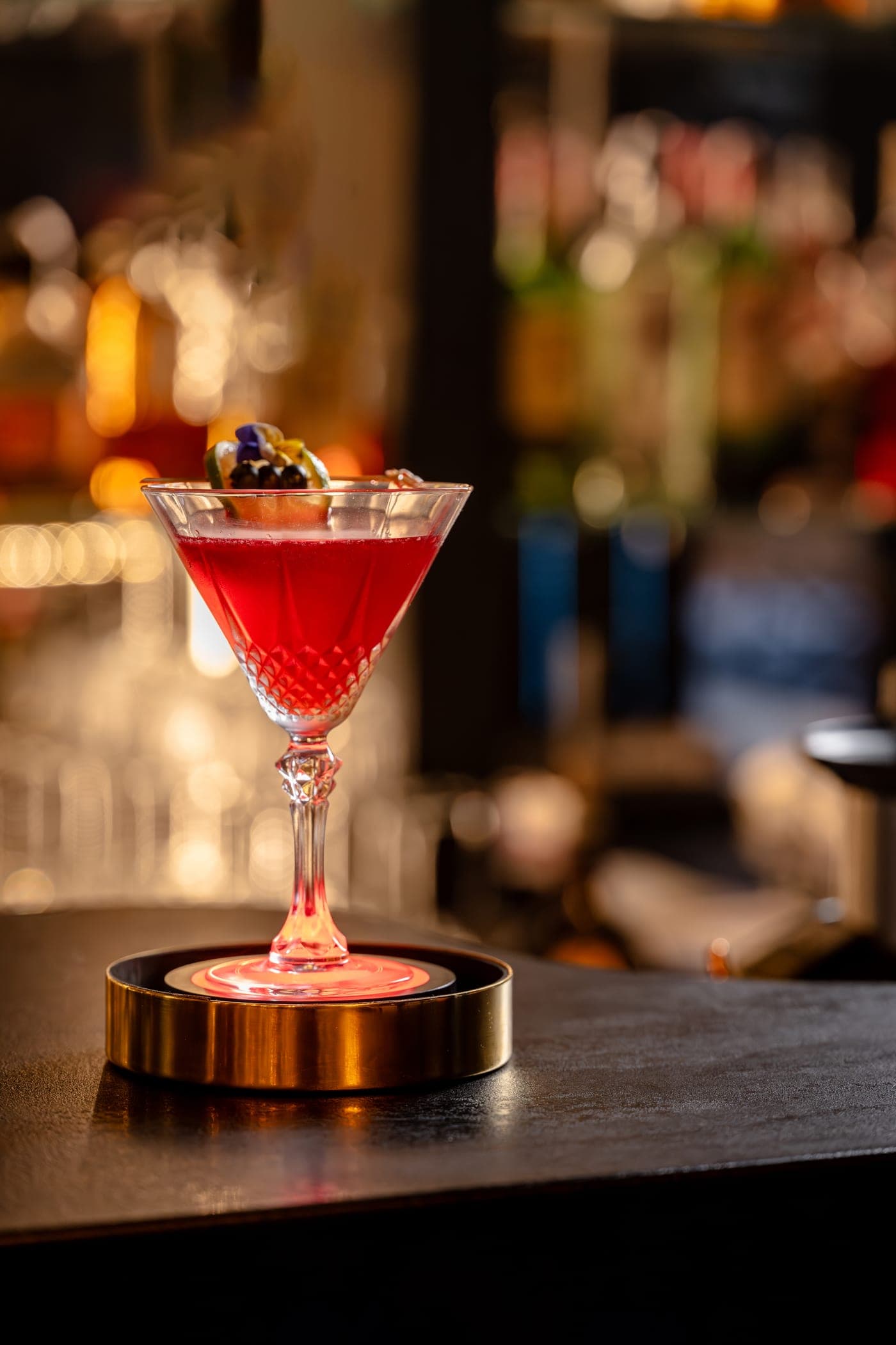 Elixirul Dragostei — cocktail rubiniu cu vișinată, merișoare, lime și afine, servit pe suport luminos auriu