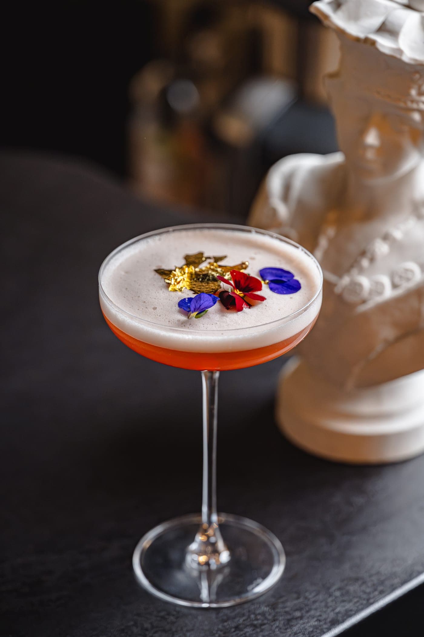Farmecele Daciei — cocktail semnătură cu spumă, foiță de aur și petale comestibile, sub bustul zeiței dace